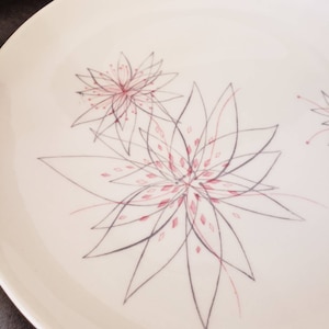 Puede incluir: Un plato de cerámica blanco con un delicado diseño floral. Las flores están dibujadas en tonos grises y rosas, creando un patrón sutil y elegante.