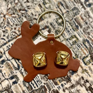 May include: A brown leather rabbit-shaped door knocker with two gold bells. The knocker has a metal ring for hanging.