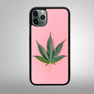 May include: A black phone case with a pink background featuring a large green cannabis leaf. The phone case has a black border and a camera lens cutout. The leaf is centered on the case.