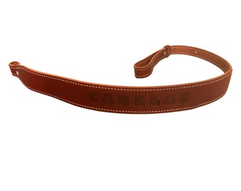 May include: A brown leather gun sling with white stitching and the word "Cabelas" embossed in the leather.