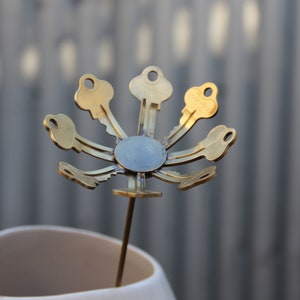 Puede incluir: Una escultura de flor de metal dorado hecha de llaves viejas. Las llaves están dispuestas en un patrón circular con las cerraduras hacia adentro. La escultura está montada en un tallo delgado de metal.