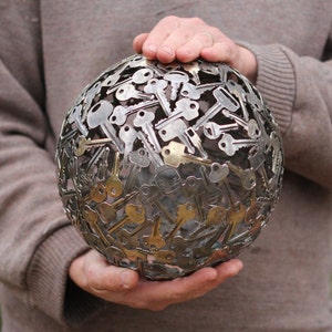 Puede incluir: Una escultura esférica hecha completamente de llaves. Las llaves están dispuestas en un patrón aleatorio, creando un diseño único e interesante. La escultura está sostenida en las manos de una persona.