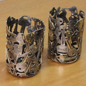 May include: Two silver and gold metal pencil holders made from old keys. The holders are cylindrical and have a rustic look.