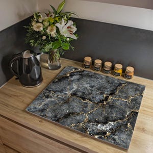 Peut inclure: Une planche à découper carrée pour cuisine présentant un motif de marbre gris foncé et noir avec des veines dorées marquées. La surface a une finition lisse et est posée sur un plan de travail en bois clair près d'une bouilloire noire.