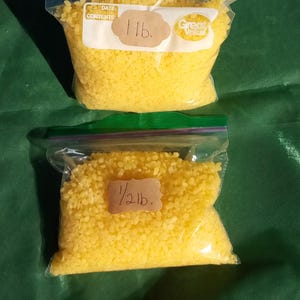 Puede incluir: Dos bolsas de plástico transparente llenas de bolitas amarillas. Una bolsa está etiquetada como "1 lb." (aproximadamente 454 gramos) y la otra como "1/2 lb." (aproximadamente 227 gramos). Las bolsas están selladas con cremallera verde, sobre una superficie verde.