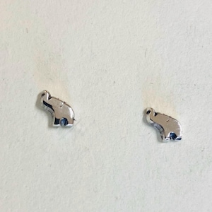 Puede incluir: Un par de pendientes de botón plateados con forma de elefante. Cada pendiente es una pequeña representación detallada de un elefante, con trompas, orejas y patas visibles. Los pendientes están sobre un fondo claro.