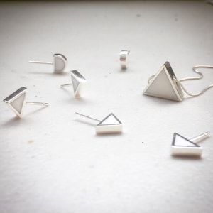 Puede incluir: Un conjunto de joyas geométricas de plata, que incluye pendientes y un collar con colgante. Los pendientes presentan un marco plateado con forma de triángulo o media luna blanca. El collar tiene un colgante triangular con el centro blanco.