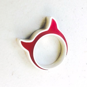 Puede incluir: Un objeto blanco y rojo en forma de anillo con un diseño único. El anillo presenta una forma redondeada con un contorno rojo y un acento rojo en la parte superior. Es probable que sea un objeto decorativo o una joya.