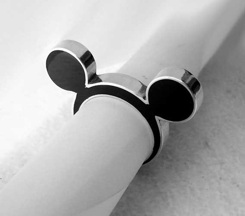May include: A silver napkin ring with a black enamel Mickey Mouse design. The ring is wrapped around a white napkin, showcasing the iconic silhouette. The design features two circular ears and a rounded head.