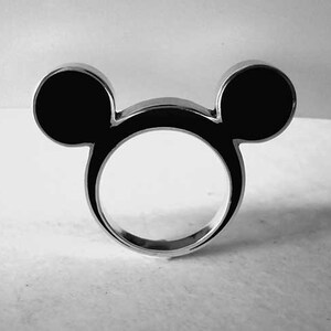 May include: A silver ring in the shape of Mickey Mouse's head. The ring has two black, circular ears and a circular band. The design is simple and modern, with a polished finish. The ring is a fun accessory.