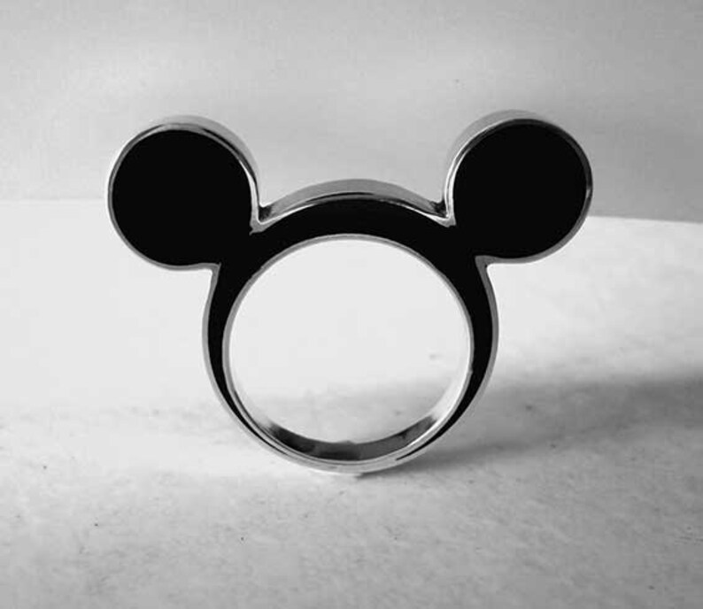 May include: A silver ring in the shape of Mickey Mouse's head. The ring has two black, circular ears and a circular band. The design is simple and modern, with a polished finish. The ring is a fun accessory.