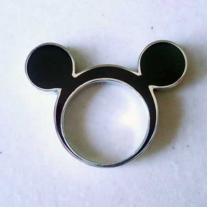 May include: A silver ring shaped like Mickey Mouse's head. The ring has two black enamel circles for ears and a circular band. A fun and playful accessory, the ring is a nod to a beloved character.