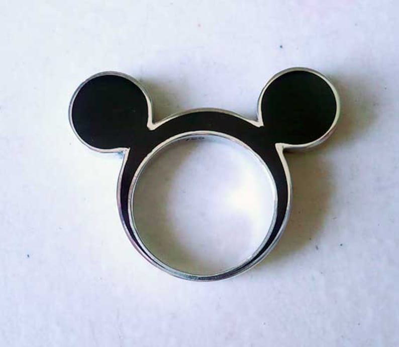 May include: A silver ring shaped like Mickey Mouse's head. The ring has two black enamel circles for ears and a circular band. A fun and playful accessory, the ring is a nod to a beloved character.