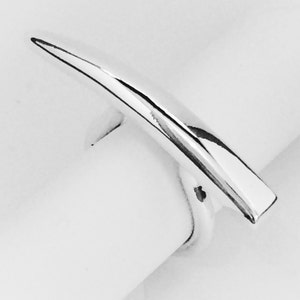 Puede incluir: Un anillo plateado con un diseño largo y puntiagudo. El anillo tiene una superficie lisa y pulida y una estética moderna y minimalista. El anillo se muestra sobre un fondo blanco.