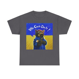 Può includere: Una maglietta grigio scuro con una grafica di un gatto che indossa un bandana rosso e una tuta blu. Il gatto è in piedi davanti a uno sfondo giallo e blu con il testo "We Can Do It!" in bianco.