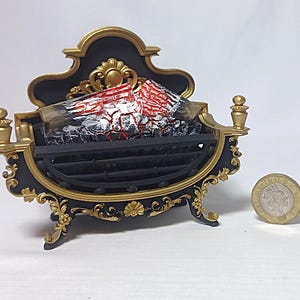 Könnte beinhalten: Ein Miniatur-Kamineinsatz in Schwarz und Gold mit kunstvollen Verzierungen, einem schwarzen Gitter und rot-weiß bemalten Holzscheiten zur Simulation eines Feuers. Eine runde silberfarbene Münze liegt daneben, um die kleine Größe zu zeigen.
