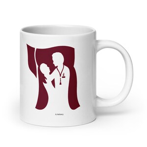 Könnte beinhalten: Weiße Keramiktasse mit einer weinroten Silhouette eines medizinischen Fachangestellten und eines Patienten. Das Design beinhaltet ein Stethoskop und den Text "A.Ashery". Die Tasse hat einen gebogenen Griff.