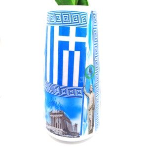 May include: A white ceramic vase with a blue and white design featuring the Greek flag, the Acropolis, and a statue of a Greek god. The vase has a Greek key border.