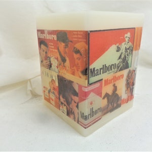 May include: A white square candle with a collage of vintage Marlboro cigarette advertisements. The ads feature cowboys, a woman, and a man smoking a cigarette. The text "Marlboro" is repeated throughout the design.