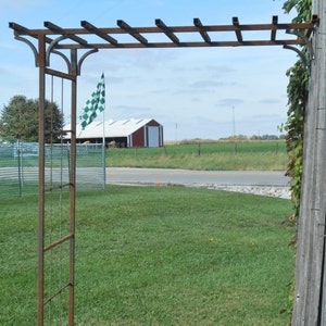 May include: A rusty metal garden archway with a trellis design. The archway is standing in a grassy field with a red barn in the background.