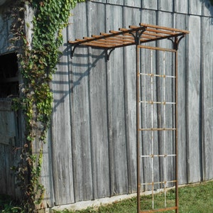 May include: A weathered, rusty metal garden arbor with a rectangular frame and a slatted top. The arbor stands on a grassy lawn against a gray wooden wall, with climbing vines adding a touch of green.