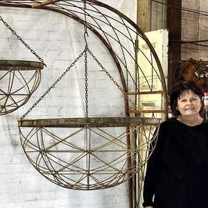 May include: Three hanging metal planters with a geometric design, suspended from a metal archway. The planters are made of a rusty metal and have a rustic look. A person is standing in front of the planters.