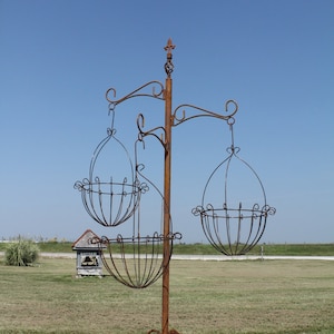 May include: A rustic, wrought-iron plant stand with three hanging baskets. The stand is brown with decorative scrollwork and a fleur-de-lis finial. The baskets are oval-shaped and designed to hold plants. The stand is outdoors against a blue sky.