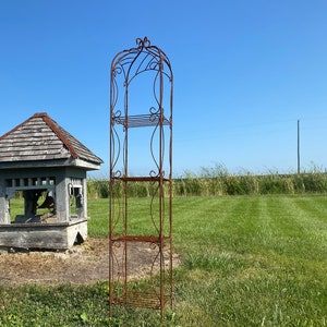 May include: A rustic wooden birdhouse with a shingled roof stands next to a tall, rusty metal plant stand with three shelves. The birdhouse is weathered and has a small opening for birds to enter. The plant stand is made of wrought iron and has a decorative design. Both structures are in a grassy field with a blue sky in the background.