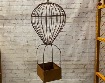 May include: A rusty metal hot air balloon planter with a square planter box hanging from the bottom. The planter is hanging from a hook on a white brick wall.