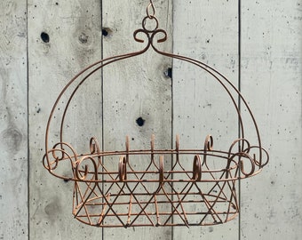 May include: A hanging, rusty-brown metal basket with a decorative, scrolled top and a diamond-patterned base. The basket is suspended by a hook and is against a weathered, light-colored wooden background.