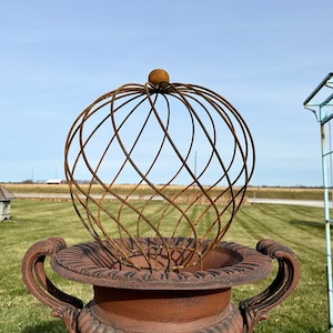 May include: A rusty metal sphere cage sits atop a brown, ornate planter with two handles. The planter is in a grassy field with a blue metal trellis in the background.