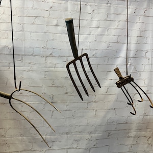 May include: Three vintage garden tools hanging from the ceiling against a white brick wall. The tools are a pitchfork, a hand rake, and a cultivator. The tools are all made of metal and have wooden handles.