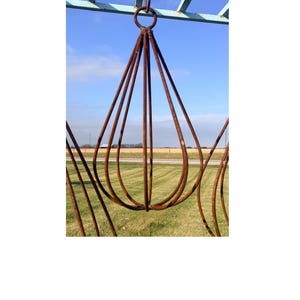 May include: A rustic, teardrop-shaped metal sculpture with a ring at the top, suspended from a light blue frame. The sculpture is made of brown, curved metal rods, set against a backdrop of a blue sky and green grass.
