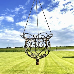May include: A hanging, ornate metal planter with a spherical base and decorative swirls. The planter is suspended by thin wires against a backdrop of a bright blue sky and green grass. A small, rustic building is visible in the background.