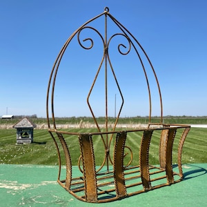 May include: A rusty brown metal hanging planter with an arched design and decorative scrollwork. The planter has a rectangular basket shape and is suspended from a single hook. The background features a blue sky and green grass.