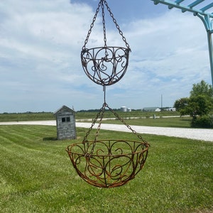 May include: Two hanging rusty metal plant baskets suspended by chains. The baskets have decorative scrollwork designs. The scene is outdoors with a blue sky, green grass, and a small wooden structure in the background.