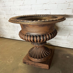 May include: A brown, weathered, cast iron planter with a fluted design and a square base. The planter is sitting on a white brick wall.