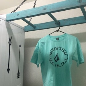 May include: A teal t-shirt with a black logo that says "Volcom Stone - Built to This" hanging on a wooden ladder in a laundry room. The t-shirt is hanging in front of a white washing machine with black arrow decals.