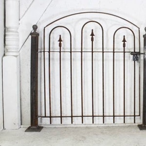 May include: A black metal gate with an arched top and decorative finials. The gate is made of vertical bars and has a latch on the right side.