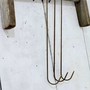 May include: Four rustic metal hooks hang from a weathered wooden beam. The hooks are made of a dark metal and have a simple, curved design.