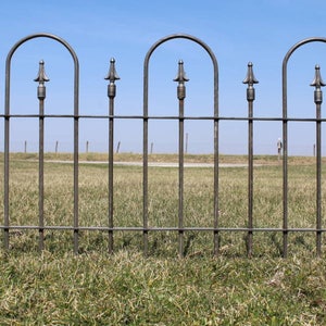 May include: A black metal fence with arched tops and spear-shaped finials. The fence is in a grassy field with a blue sky in the background.