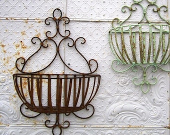 May include: Two decorative metal wall planters. One is a rusty brown color, and the other is a faded green. Both have a basket shape with vertical bars and ornate scrollwork. They are mounted on a white textured wall.