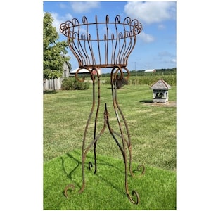 May include: A tall, ornate, rusty metal plant stand with a circular basket-like top. The stand has a three-legged base with decorative scrollwork and a pointed accent. The stand is outdoors on a grassy lawn.