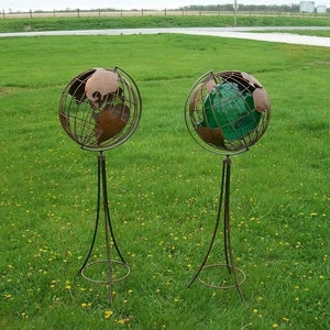 May include: Two metal globes on stands, one with a brown globe and the other with a green globe. The globes are on a grassy lawn.