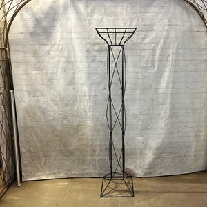 May include: A tall, black metal plant stand with a geometric design. The stand features a square base, a slender, twisted central column, and a square top with a grid pattern. The stand is designed to hold a potted plant.