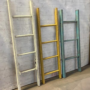 May include: Four wooden ladders painted in different colors. The ladders are leaning against a white brick wall. The ladders are white, yellow, blue, and turquoise.