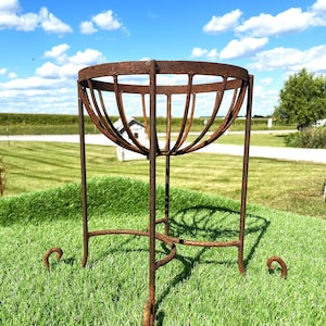 May include: A rusty metal plant stand with a circular top and curved vertical supports. The stand has three legs with curled feet and is set on a grassy lawn under a blue sky with clouds. The stand is designed to hold a plant pot.