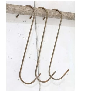 May include: Three rustic metal hooks hang from a wooden beam. The hooks are made of a dark metal with a slightly rusted finish. They are all the same size and shape, with a long, curved arm and a sharp point at the end.