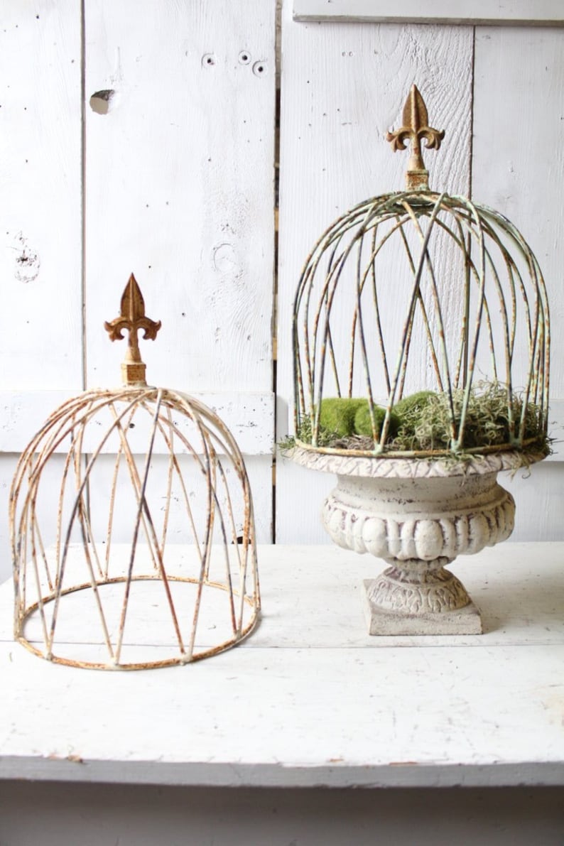May include: Two white metal plant cages with gold finials. The larger cage is on a white pedestal and has green moss inside. The smaller cage is on a white wooden surface.