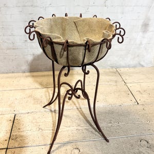 May include: A brown metal plant stand featuring a circular frame with decorative scrollwork accents and a tall, slender base. The bowl-shaped top is lined with a thick, beige felt fabric insert that conforms to the curved wire structure.